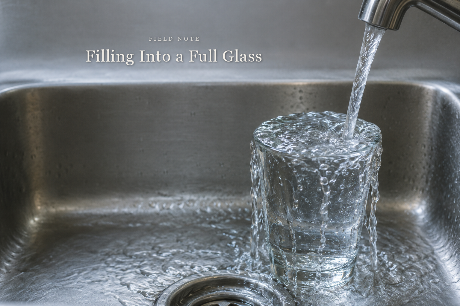 A clear drinking glass standing in a stainless steel kitchen sink, filled to the brim, with a thin stream of water running into it from a tap and spilling evenly over the rim. The article title is set into the quiet space above.