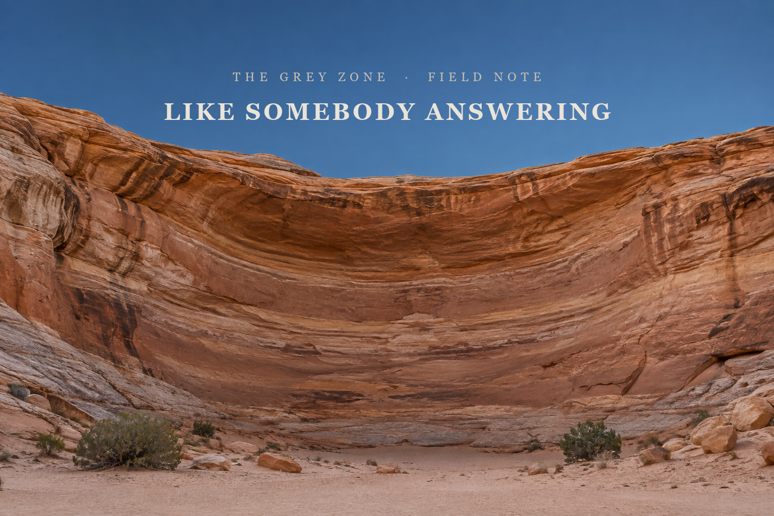 A wide, curved alcove in a layered sandstone canyon wall, seen from the canyon floor in soft early light, with pale sand and a few low shrubs at its foot and a clear, deep blue sky above. The article title is set in the sky.